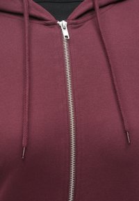 Sweat à capuche zippé bordeaux avec une fermeture éclair argentée. Il possède une capuche à cordon et une texture côtelée autour du cou. Tissu doux.