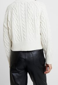 Weißer Kabelstrickpullover mit einem cropped Design und geripptem Saum, kombiniert mit hochgeschnittenen schwarzen Lederhosen. Strukturierte und gemütliche Optik.
