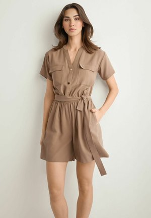 Kobieta o ciemnych włosach do ramion, ubrana w beżowy krótki kombinezon z paskiem i przednimi kieszeniami, stojąca na jednolitym jasnym tle.