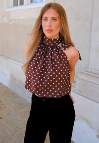 Γυναίκα με μακριά ανοιχτοκάστανα μαλλιά, που φοράει καφέ αμάνικο τοπ με πουά και μαύρο παντελόνι, στέκεται μπροστά σε έναν ανοιχτόχρωμο πέτρινο τοίχο έξω.