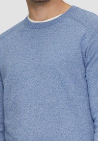 Jasnoniebieski dziany sweter wykonany z teksturowanej tkaniny, z okrągłym dekoltem i rękawami raglanowymi. Zbliżenie ukazuje miękkość materiału.