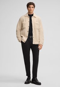 Beige fleece jacket with two front pockets, button closure, and rounded collar, paired with black tapered pants and black shoes.