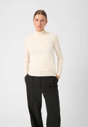 Maglione a collo alto beige con una vestibilità morbida e aderente e maniche lunghe, abbinato a pantaloni neri larghi, che accentuano uno stile minimalista.