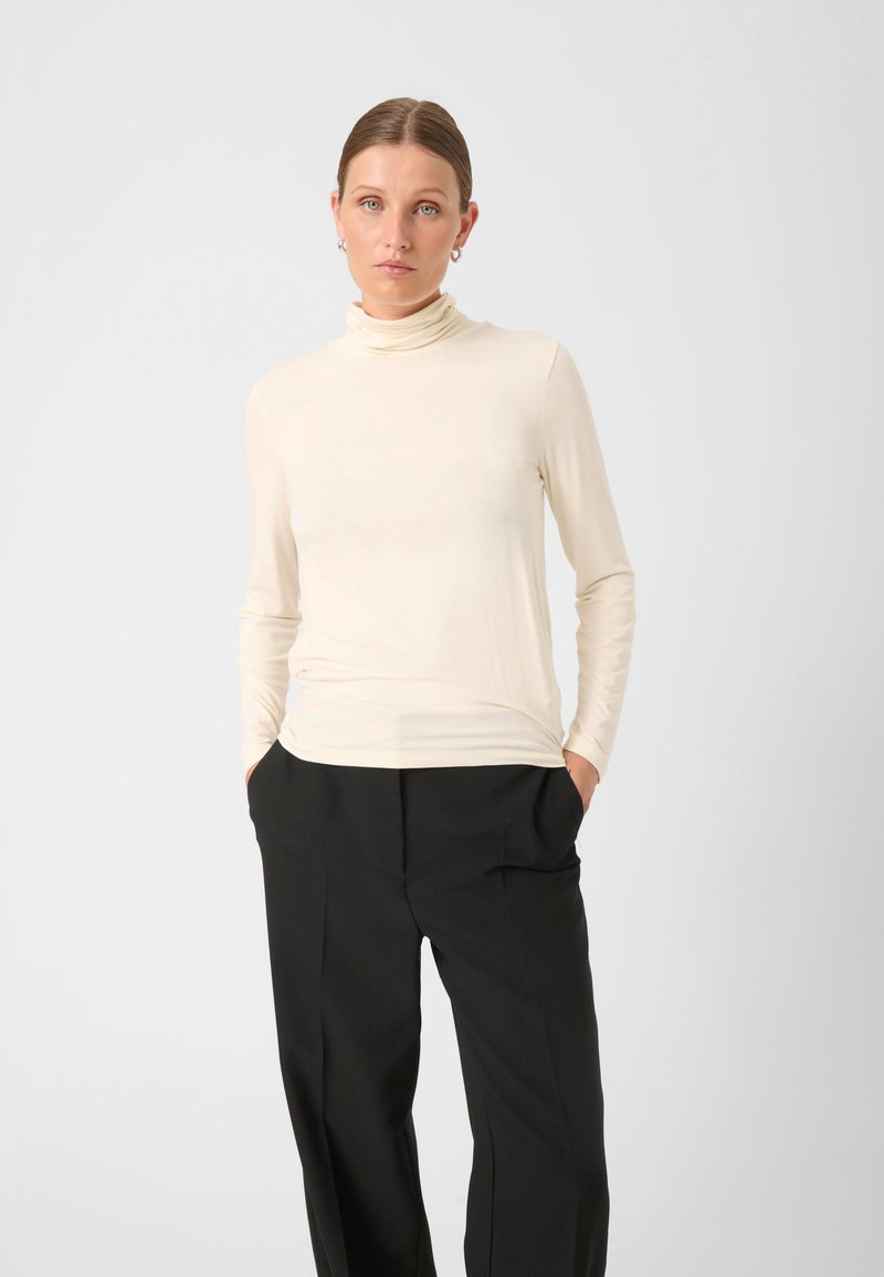 Cremfarbener Rollkragenpullover in einem glatten, figurbetonten Design mit langen Ärmeln, kombiniert mit lockeren, schwarzen Hosen, die einen minimalistischen Stil betonen.