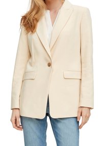 Femme portant un blazer crème à bouton unique par-dessus un haut blanc et un jean bleu, vue des épaules jusqu'aux hanches.
