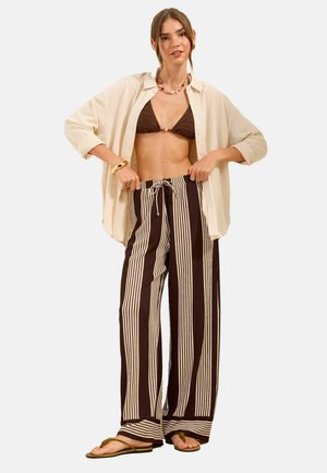 Kvinde iført en beige åben skjorte, brun bikini-top, stripede bukser med vide ben, sandaler og smykker, stående med hænderne og justerer bukselinningen.