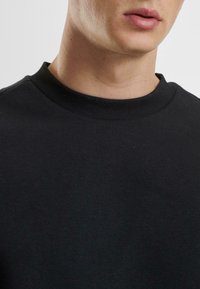 Schwarzer Rundhalsausschnitt-Sweatshirt aus weichem Stoff. Verfügt über einen gerippten Kragen und Ärmelabschlüsse. Nahaufnahme des Kragens und des oberen Brustbereichs.
