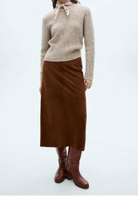 Brauner Wildleder-Midirock, kombiniert mit einem beigen strukturierten Pullover, akzentuiert durch einen hellen Schal und kniehohen braunen Stiefeln. Glatte Textur.