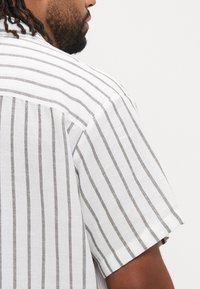 Πουκάμισο με κοντά μανίκια σε λευκό χρώμα με κάθετες καφέ ρίγες. Διαθέτει υφασμάτινη υφή και άνετη εφαρμογή, αναδεικνύοντας ένα casual σχέδιο.