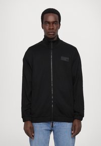 Giacca nera con zip, colletto alto, tessuto testurizzato e due tasche laterali. Presenta tre strisce nere lungo ciascun manica e un distintivo.