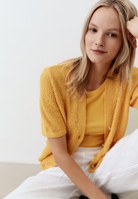 Gelber, strukturierten Cardigan mit kurzen Ärmeln, kombiniert mit einem passenden gerippten Shirt. Das Modell trägt hellfarbene Hosen und sitzt vor einem neutralen Hintergrund.