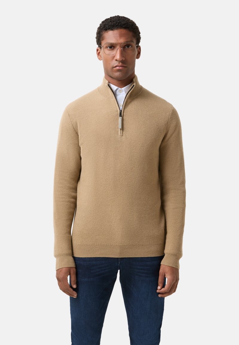Beige pletený svetřík s vysokým límcem a čtvrtovým zipem. Má dlouhé rukávy a přiléhavý střih, doplněný tmavými džínami.