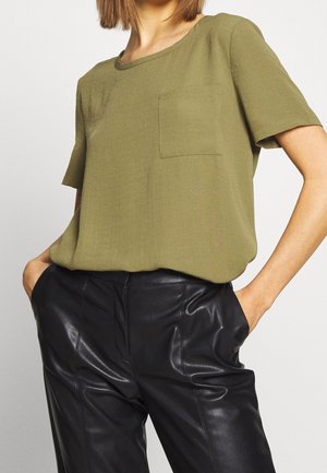 Mujer con camisa de manga corta color verde oliva con bolsillo en el pecho, metida dentro de pantalones de cuero negros de talle alto, con las manos en los bolsillos.