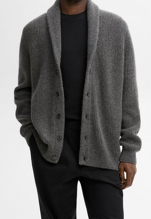 Homme portant un cardigan côtelé gris foncé avec des boutons, une chemise noire en dessous et un pantalon noir, debout les mains dans les poches.