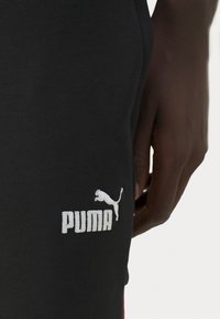 Pantaloni sportivi neri con logo Puma ricamato bianco sulla coscia. Il tessuto appare liscio con una leggera elasticità.