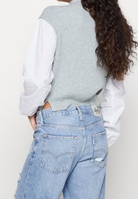 Maglione senza maniche a coste grigio chiaro abbinato a una camicia bianca a maniche lunghe e jeans azzurri chiari strappati, che mette in mostra i dettagli sul retro.