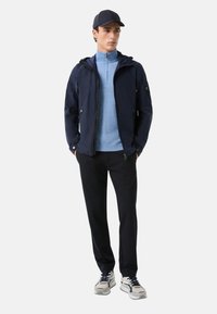 Marineblaues Jackett mit Reißverschlusstaschen über einer hellblauen Zwischenschicht, kombiniert mit schwarzen Hosen und hellen Sneakers, die eine Schnürung und strukturierte Details aufweisen.