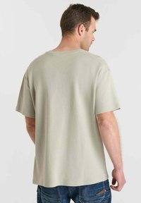 Lichtgrijs T-shirt met korte mouwen van een gestructureerde stof, relaxte pasvorm en een ronde hals, gezien van achteren. Spijkerbroek is zichtbaar.