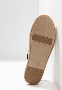 Semelle d'une chaussure espadrille beige avec grip en caoutchouc texturé, côtés en jute tissé visibles, et logo "TOMS" embossé sur la semelle.