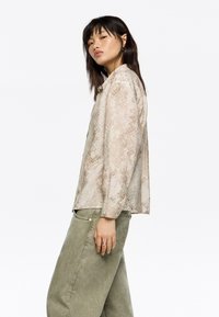 Camisa de manga larga en un beige claro con un estampado de serpiente. Corte holgado, textura suave, cierre de botones, combinada con pantalones verde olivo.