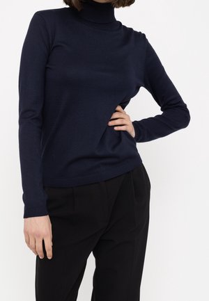 Femme portant un pull à col roulé bleu marine et un pantalon noir, une main sur la hanche et l'autre détendue le long du corps.