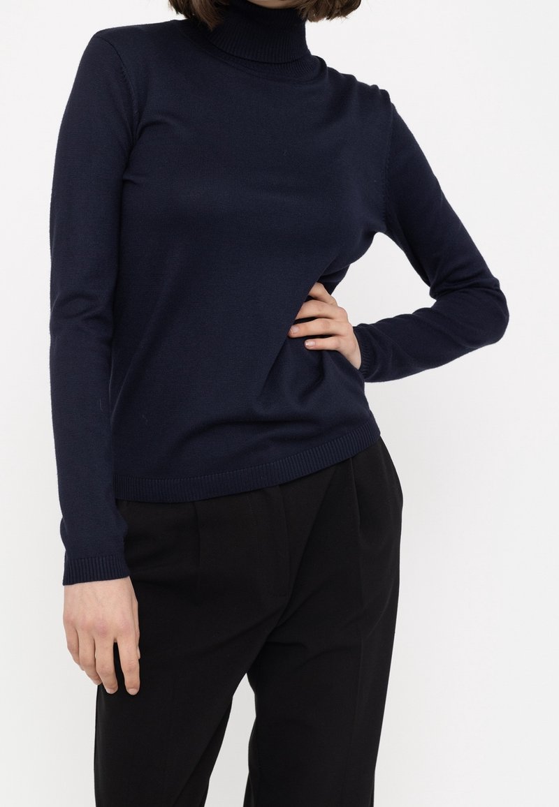 Femme portant un pull à col roulé bleu marine et un pantalon noir, une main sur la hanche et l'autre détendue le long du corps.