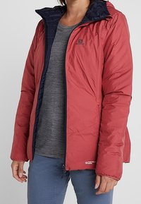 Rote isolierte Jacke mit navyfarbenem Futter. Verfügt über eine Kapuze, einen Reißverschluss und Logodetails. Getragen über einem grauen Shirt und blauen Hosen.