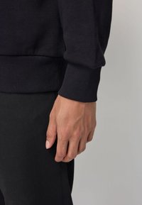 Felpa nera con polsini a coste, con una mano appoggiata su jeans neri. Focus sulla texture del tessuto e sulle linee pulite del design del capo.