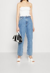 Weißes trägerloses Oberteil, blaue hochtaillierte Jeans, weiße Plateausandalen und eine Handtasche in Schwarz und Weiß mit abgerundeter Form.