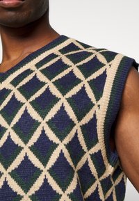 Strickweste ohne Ärmel mit einem geometrischen Rautenmuster in Marineblau, Creme und Grün. Texturiertes Strickmaterial mit runder Ausschnittform und subtilen Rippungen.
