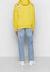 Person som bär en gul hoodie med blå, vit och röda Nike-loggor på ryggen, ljusblå jeans och vita sneakers, står på en grå golv.