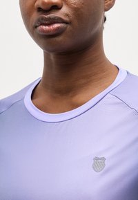 Close-up af en person iført en lyse lilla T-shirt med rund hals og et lille K-Swiss-logo på brystet.