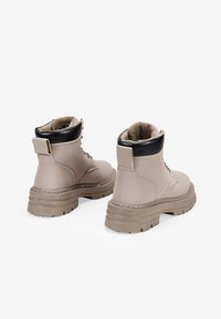 Bottines beige avec un col rembourré noir, un devant à lacets, une semelle en caoutchouc et une doublure en fausse fourrure. Dispose d'une languette décorative à l'arrière et d'accents cousus.