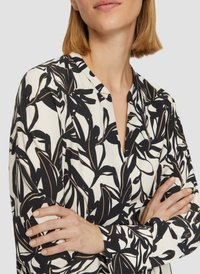 Lange mouwen blouse van witte stof met een zwart en gouden bladerenpatroon, met een kleine halslijn en een enigszins gestructureerd oppervlak.