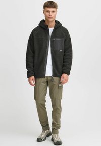 Hoodie en polaire noire avec une poche zippée sur la poitrine, associé à un pantalon cargo olive et des chaussures de randonnée grises. Design simple et tissu texturé.