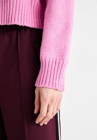 Stickad rosa tröja med ribbade muddar, ihop med mörk burgundys färgade byxor. Handen visar en liten tatuering nära handleden.