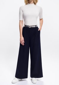 Femme debout portant un haut blanc à manches courtes, un pantalon large navy à rayures avec ceinture, et des baskets blanches sur un fond blanc.