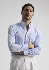 Camisa de vestir a rayas en azul claro y blanco con cuello puntiagudo, botonadura frontal y un bolsillo en el pecho. La tela parece suave y nítida.