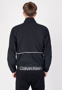 Giacca a vento nera con colletto alto, polsini elasticizzati e una striscia orizzontale bianca in vita. Presenta un logo Calvin Klein sulla vita.