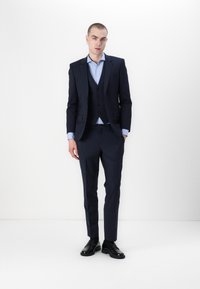 Marineblauwe pak met een getailleerde blazer, bijpassend vest en broek. Lichtblauw overhemdd onder. Zwarte schoenen maken de look af.