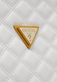Hvid quilted syntetisk håndtaske med et trekantet guldaccent og en hvid indsats, der står "GUESS U.S.A." samt et spørgsmålstegn.