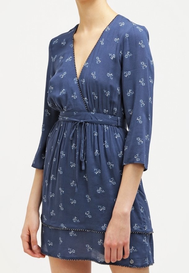 Femme portant une robe portefeuille bleue à fleurs avec des manches trois-quarts et une ceinture à la taille, debout devant un fond uni.