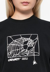 Black cotton t-shirt featuring a white spider web graphic, including a small spider detail, and "CARHARTT WIP THEORY & PRACTICE" text.