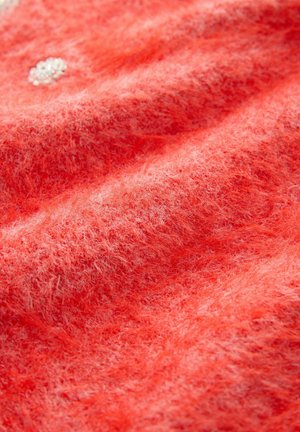 Zblízka na mäkkú, nadýchanú koralovú tkaninu s viditeľnými vláknami a malým súborom bielych korálok prišitých v blízkosti horného ľavého rohu.