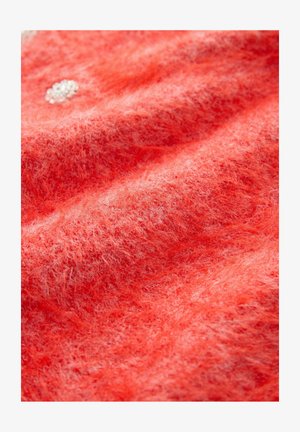 Close-up van zacht, fluffig koraalstoffen met zichtbare vezels en een kleine groep witte kralen die aan de bovenlinkerzijde zijn genaaid.