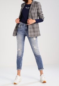 Grå rutig blazer över en marinblå topp, kombinerad med slitna ljusblå jeans och vita sneakers med tanfärgade sulor. Avslappnad och modern outfit.