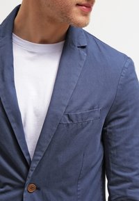 Námořnický modrý lněný blazer s jednobuttonovým zapínáním, klopovými límci a náprsní kapsou, položený přes bílé tričko. Hladká textura.