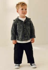 Veste en denim gris avec capuche, boutons sur le devant et motifs de smileys. T-shirt blanc en dessous, associé à un pantalon cargo noir ample et des sneakers.