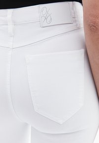 Los pantalones vaqueros de mezclilla blanca presentan un diseño ajustado, un bolsillo trasero con costuras reforzadas y una etiqueta de marca en la cintura.