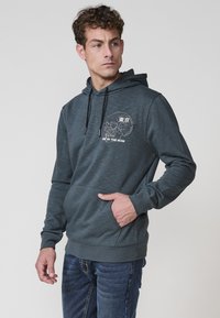 Sötét szürke kapucnis pulóver, puha anyagból készült, elülső zsebbel. Nyomtatott dizájnnal rendelkezik, szöveggel és kör alakú logóval. Állítható zsinóros kapucni.